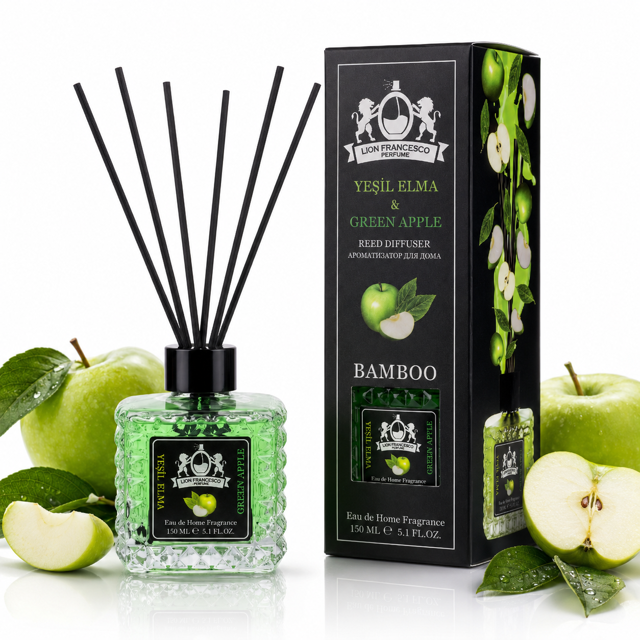A reed diffuser bottle with black reeds and a green liquid, shown next to its branded box. The scene is styled with whole and sliced green apples and leaves on a clean white background.