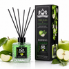 A reed diffuser bottle with black reeds and a green liquid, shown next to its branded box. The scene is styled with whole and sliced green apples and leaves on a clean white background.