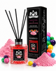 A reed diffuser bottle filled with pink sugar-like beads stands beside its black branded box, surrounded by pink powder piles, colorful candy-like balls, and diffuser reeds on a clean white background.